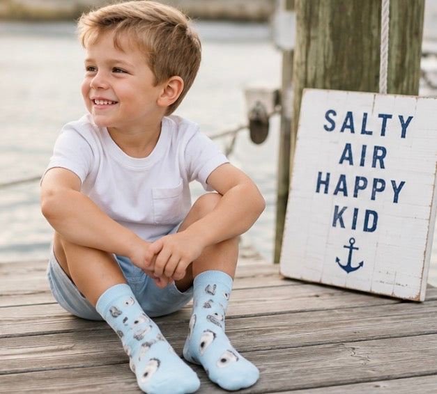 Boy wearing baby blue Bonfolk oyster socks sitting on a dock next to a "Salty Air Happy Kid" sign