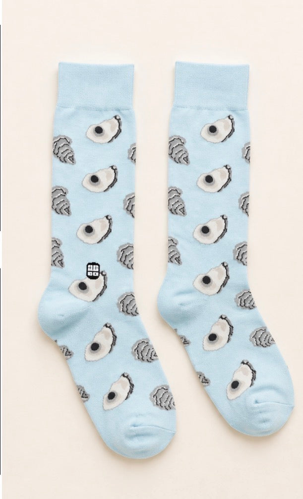 A pair of light blue crew socks with a repeating pattern of cartoon avocado faces and small gray striped figures, displayed flat against a light beige background.