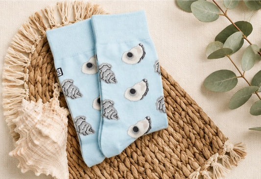 A pair of light blue crew socks with a pattern of gray and white seashells, displayed on a woven straw mat with a seashell and eucalyptus leaves.