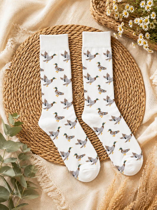 A pair of white crew socks with an all-over print of mallards, styled in a flat lay on a round woven placemat over a beige fabric background with dried flowers and greenery.